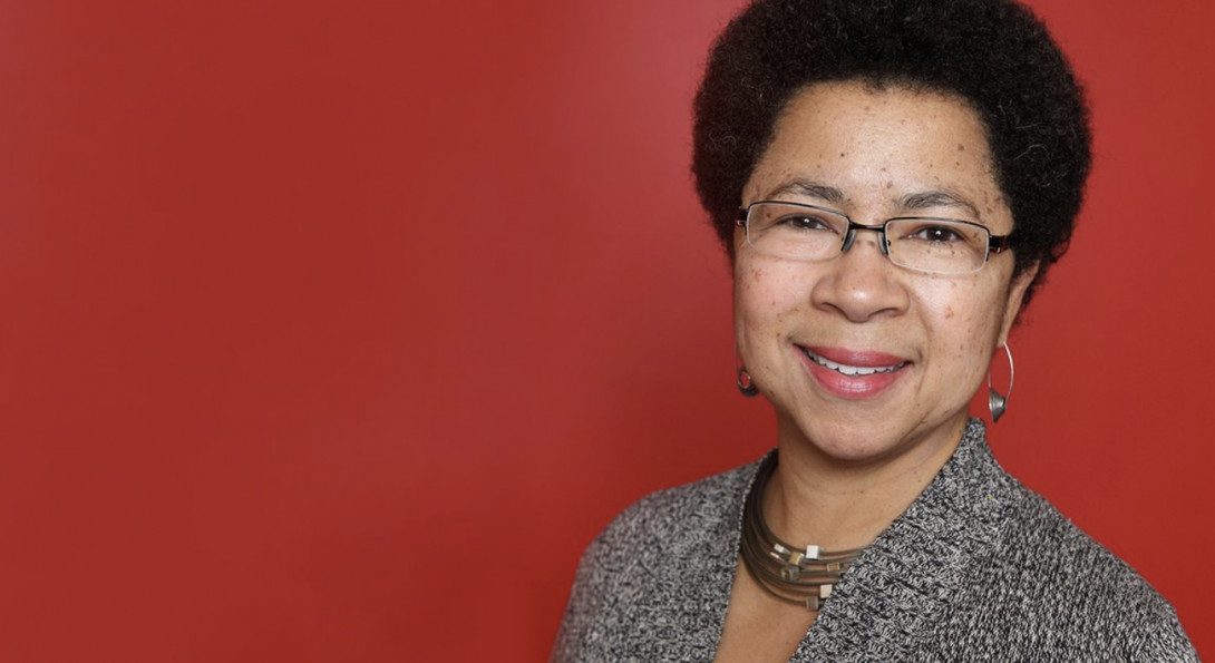 Dr. Barbara Ransby wearing a grey knit top and gold necklace stands before a red background